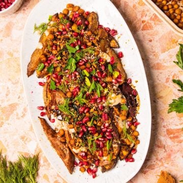 Roasted eggplant on a white platter topped with chickpeas, herbs, pomegranate seeds, and sauce-perfect alongside roasted sweet potatoes with tahini for a vibrant, flavorful spread.