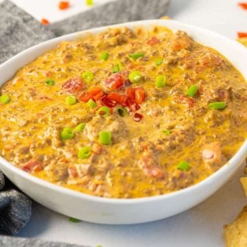 A bowl of cheesy dip with ground meat, topped with diced tomatoes and green onions, next to tortilla chips.