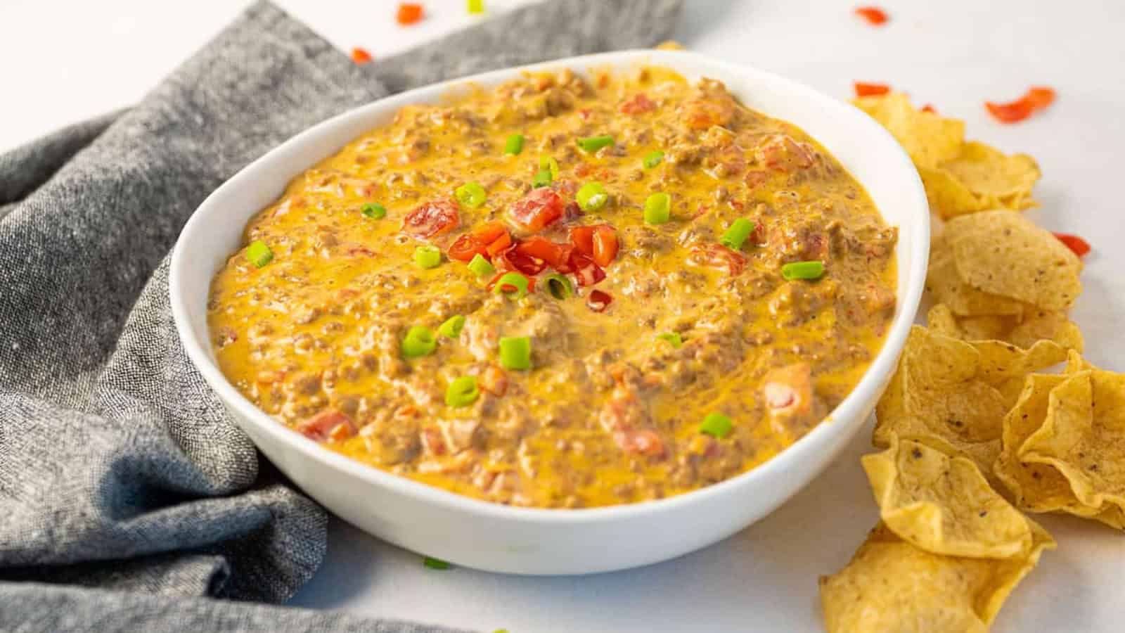 A bowl of cheesy dip with ground meat, topped with diced tomatoes and green onions, next to tortilla chips.