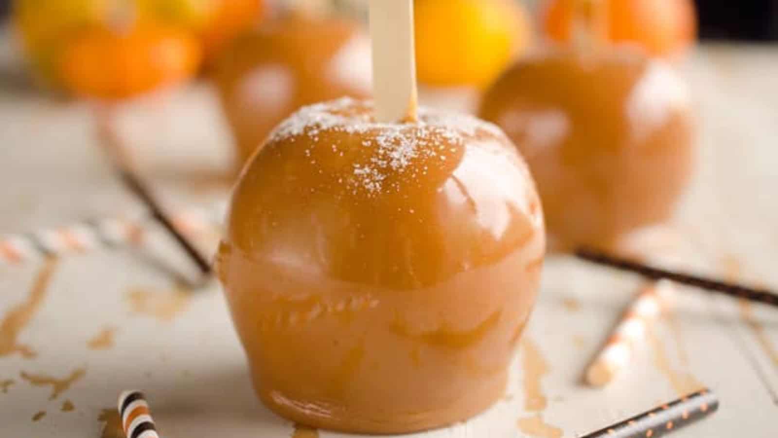 Close-up of caramel-coated apple with a stick, sprinkled with sugar, and blurred apples in the background.