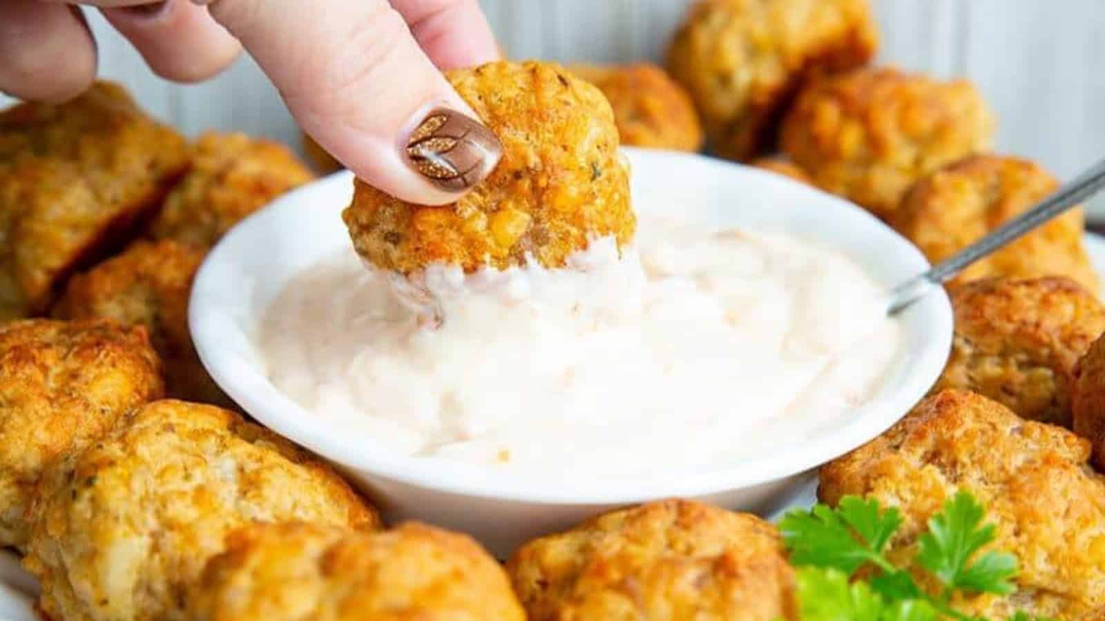 Hand dipping a cheesy sausage ball into a bowl of creamy dip, surrounded by more sausage balls.