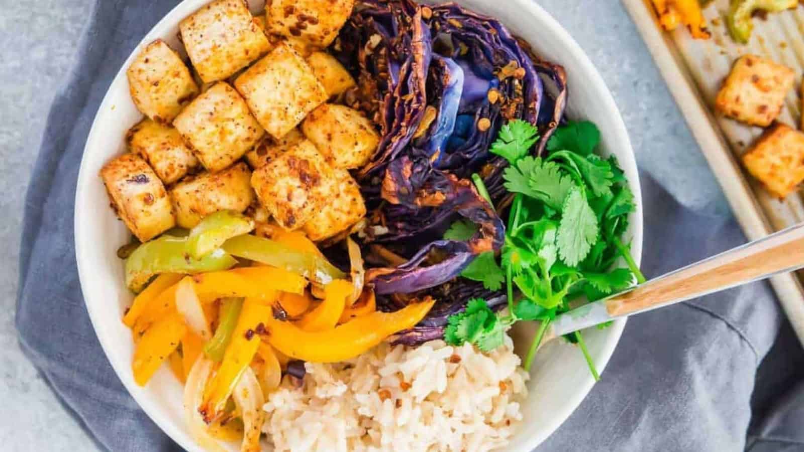 Bowl with brown rice, tofu cubes, purple cabbage, sautéed peppers, and fresh cilantro, with a fork.