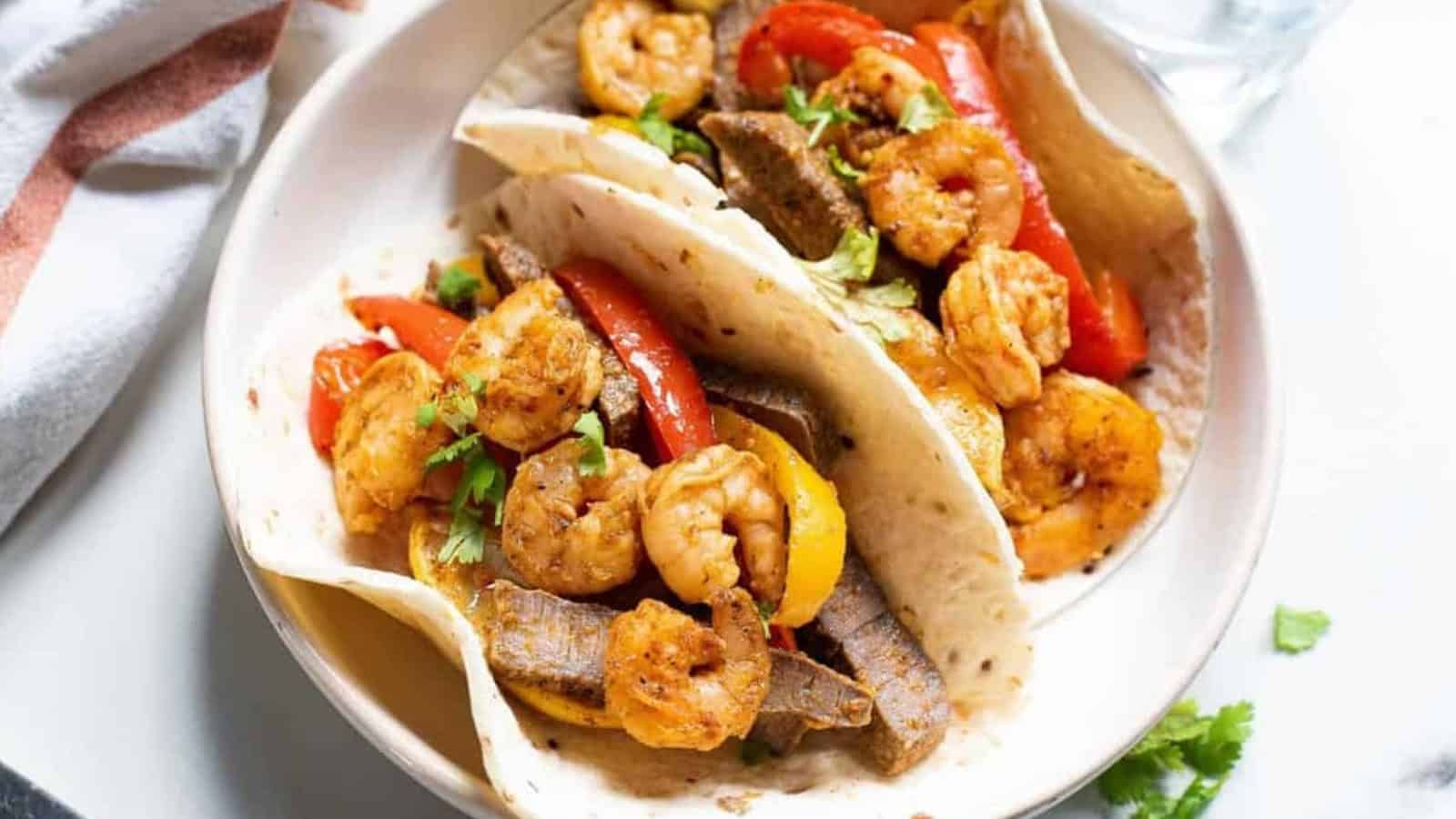 Two tortillas filled with shrimp, beef strips, bell peppers, and cilantro on a white plate.