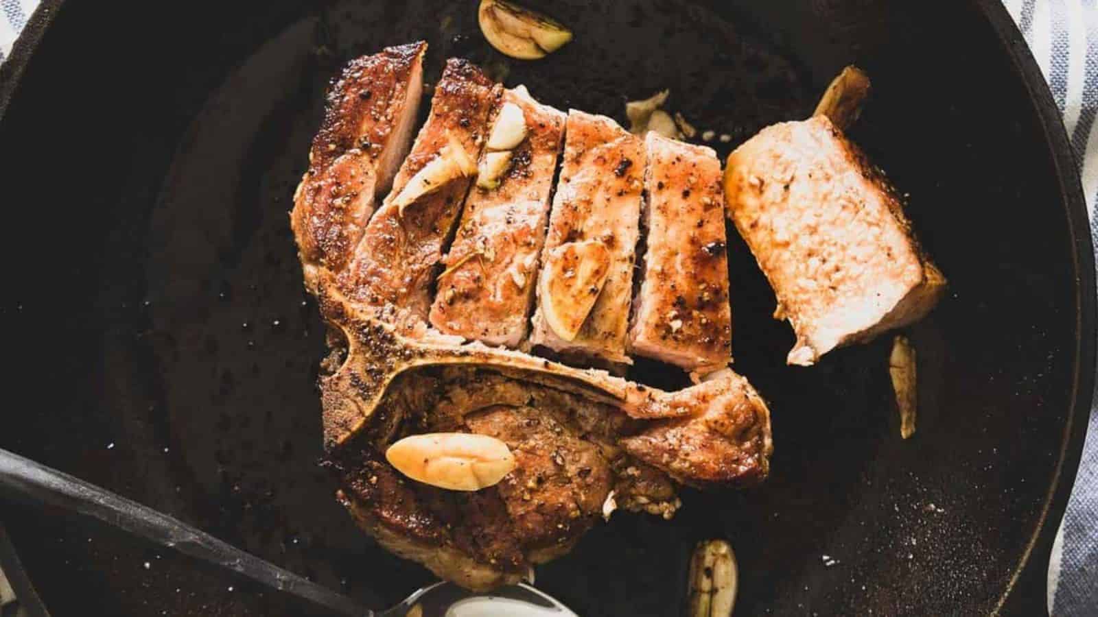 Sliced grilled pork chop with garlic pieces on a dark skillet, partially cut with a spoon nearby.