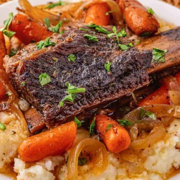 Braised short rib with carrots and onions served over mashed potatoes, garnished with chopped parsley.