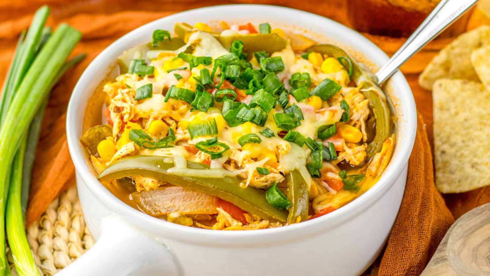 A white bowl filled with a mixture of shredded chicken, green bell peppers, corn, melted cheese, and chopped green onions with a spoon inside the bowl. Tortilla chips are on the side.