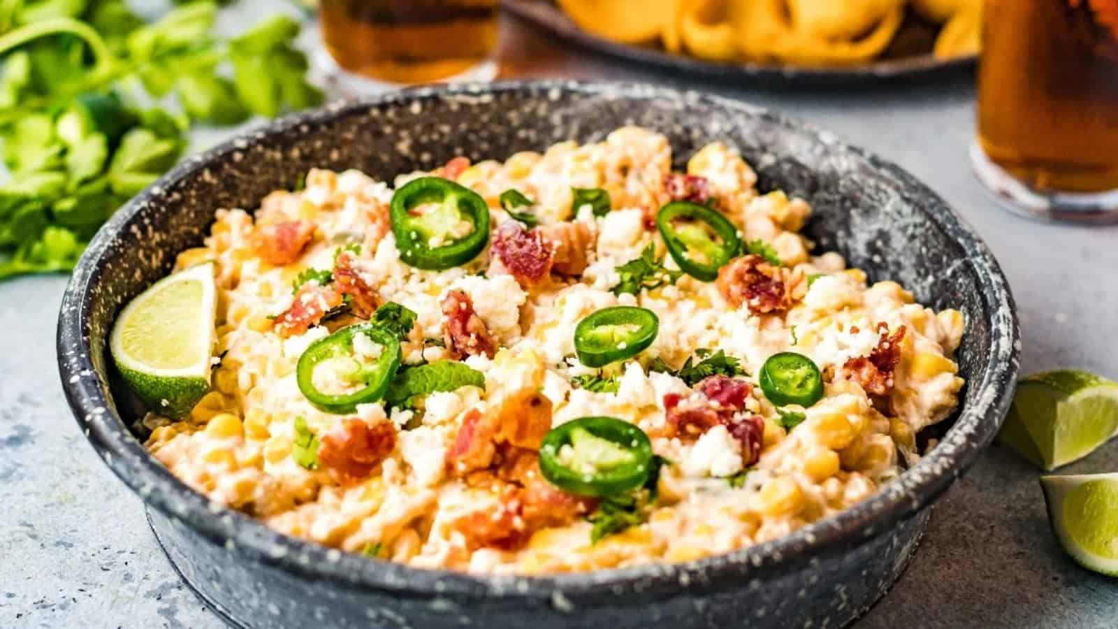 Creamy corn salad topped with sliced jalapeños, bacon, cheese, and herbs, served with lime wedges in a black bowl.