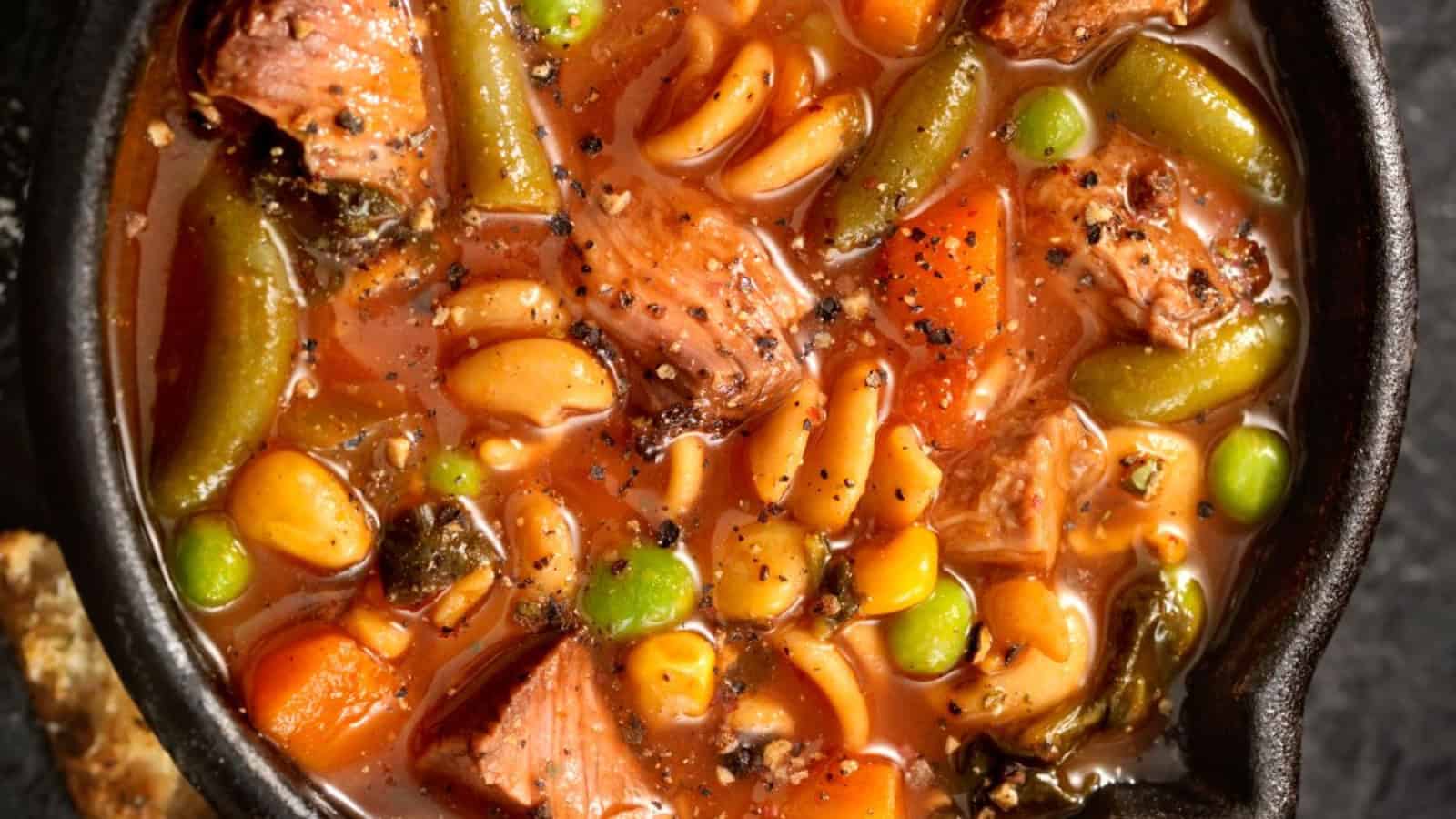 Bowl of hearty beef and vegetable soup with pasta, beans, peas, carrots, green beans, and cracked black pepper.