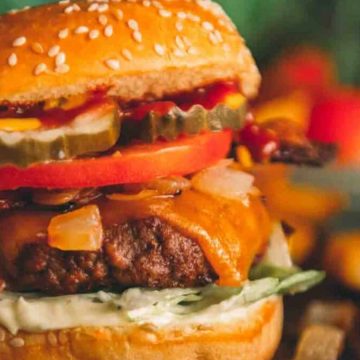 Close-up of a cheeseburger with lettuce, tomato, pickles, onions, bacon, and melted cheese on a sesame bun.