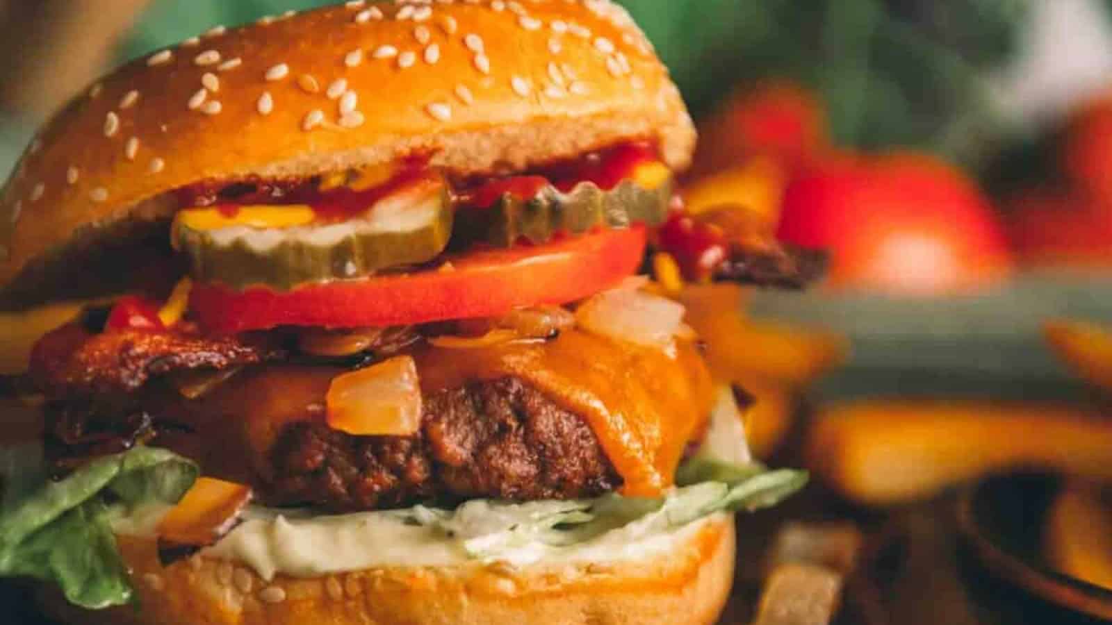 Close-up of a cheeseburger with lettuce, tomato, pickles, onions, bacon, and melted cheese on a sesame bun.