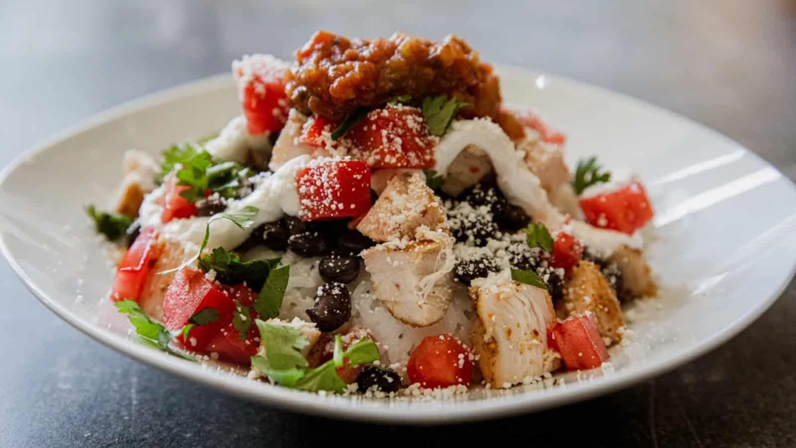 A plate of chicken, black beans, tomatoes, cheese, salsa, and cilantro served over rice with a drizzle of sour cream.