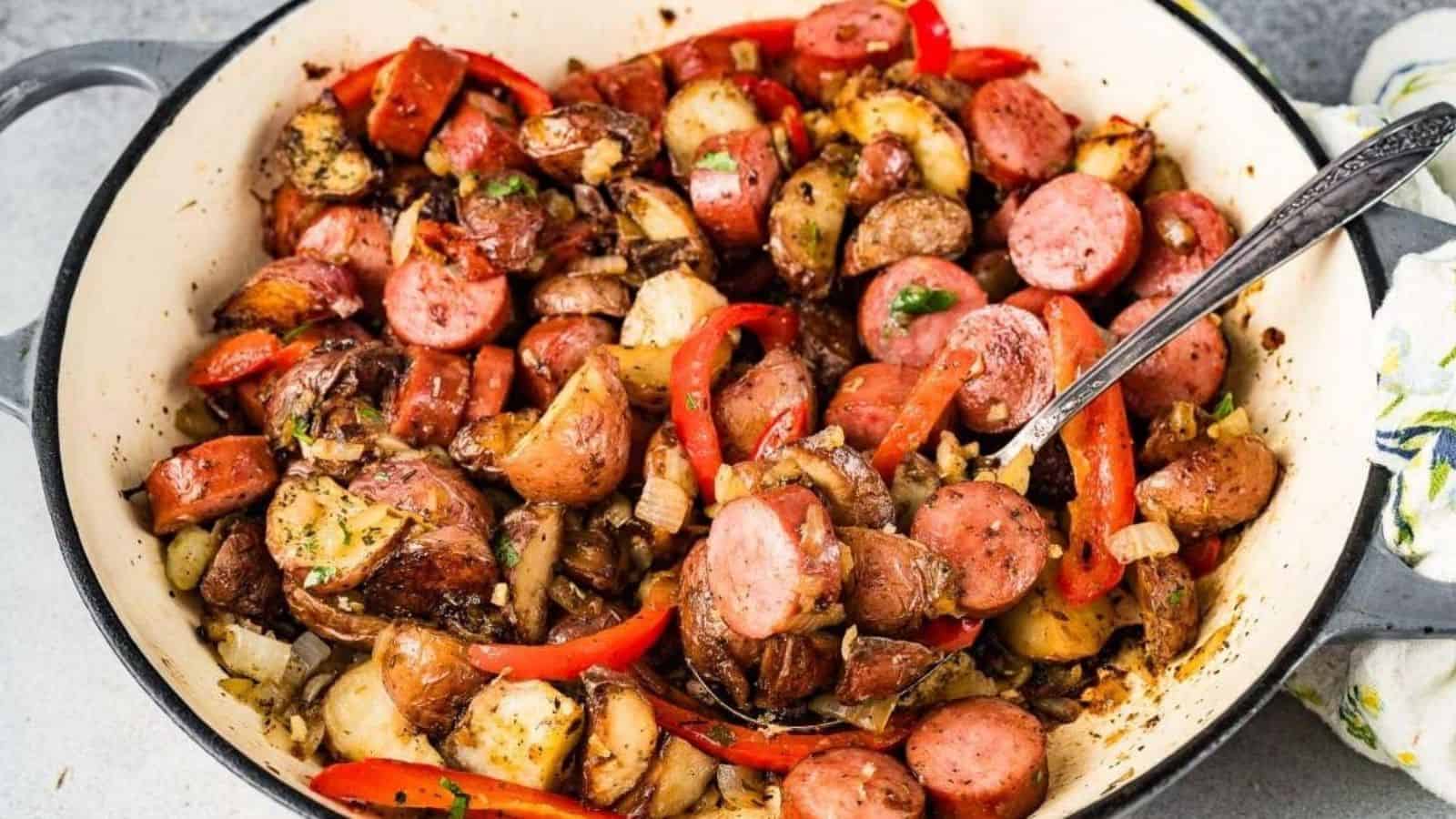 Sausage, potatoes, onions, and red bell peppers cooked together in a large skillet with a serving spoon.