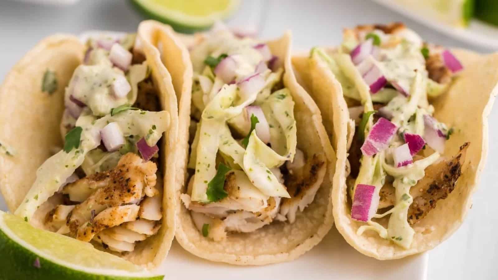Three fish tacos topped with cabbage, diced red onions, and creamy sauce, with lime wedges on the side.