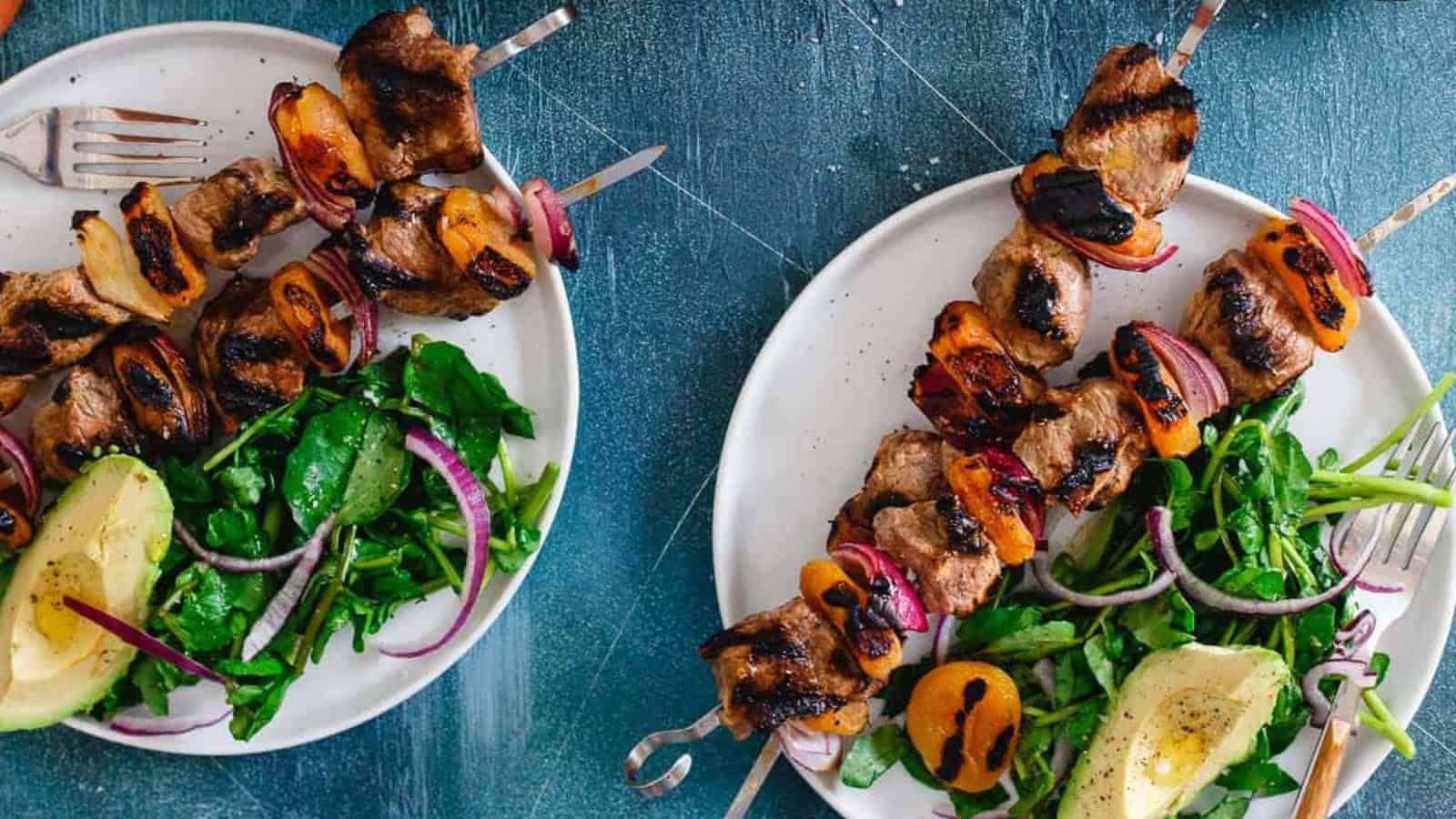 Two plates with grilled meat skewers, salad greens, red onion, and avocado halves on a blue surface.