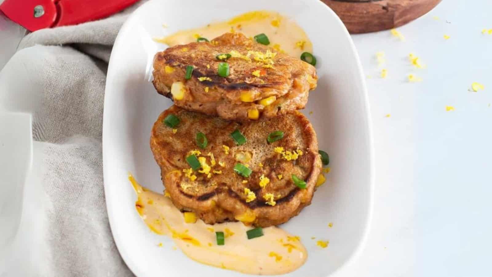 Two corn fritters on a white plate, garnished with green onions and sauce, sprinkled with lemon zest.