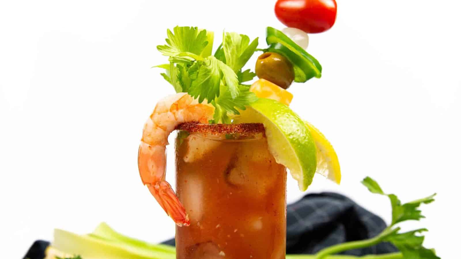 A Bloody Mary garnished with celery, shrimp, lime, lemon, and skewered vegetables against a white background.