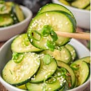 Chopsticks holding cucumber slices with sesame seeds and green onions over a bowl of cucumber salad.