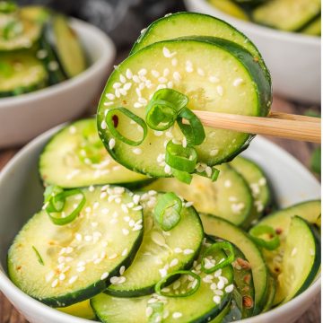 Chopsticks holding cucumber slices with sesame seeds and green onions over a bowl of cucumber salad.