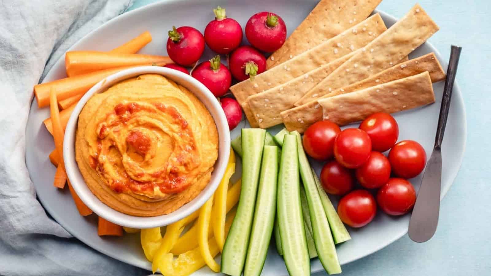 A platter with hummus, carrot sticks, radishes, crackers, cucumber sticks, yellow peppers, and cherry tomatoes.