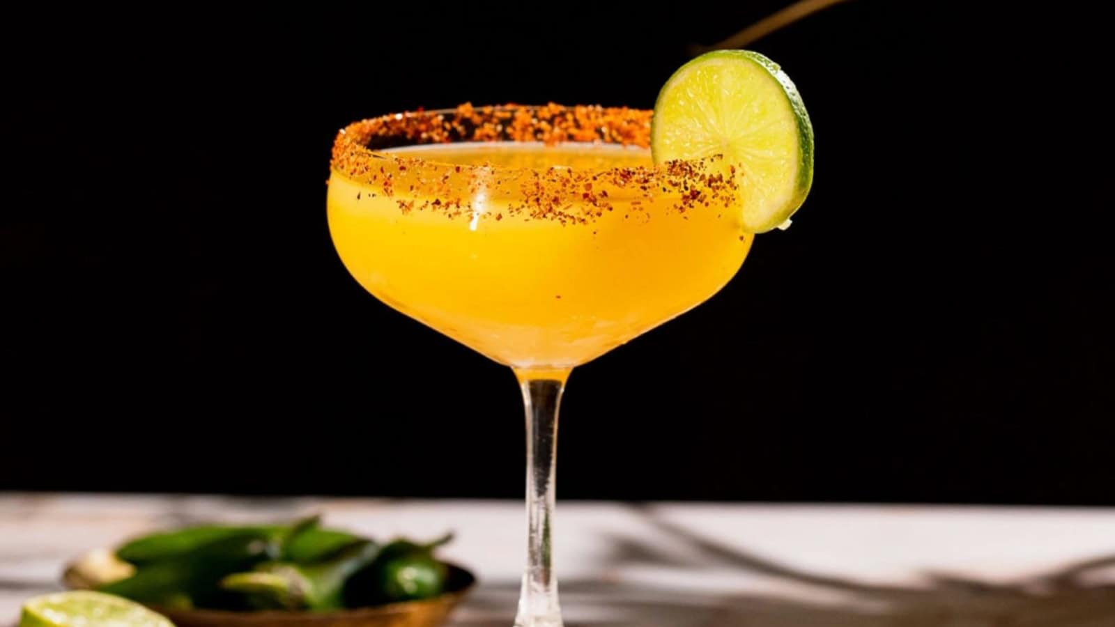 Yellow cocktail in a glass with a chili rim and a lime slice garnish, with limes and peppers nearby.