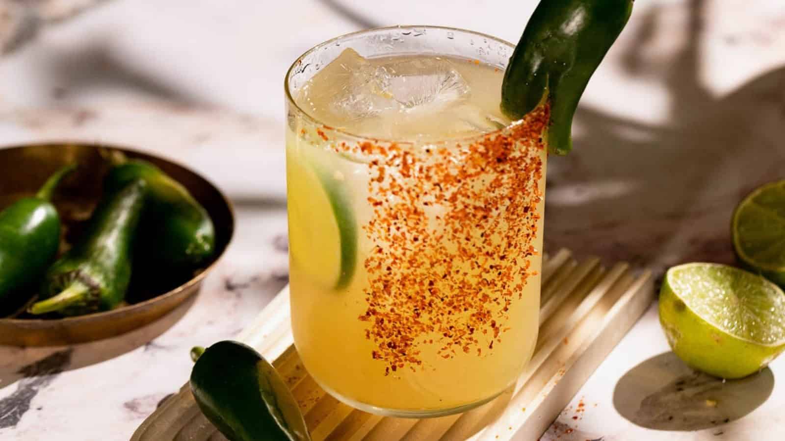 A spicy margarita with chili powder rim, lime wedges, ice, and a jalapeño garnish, on a white surface.
