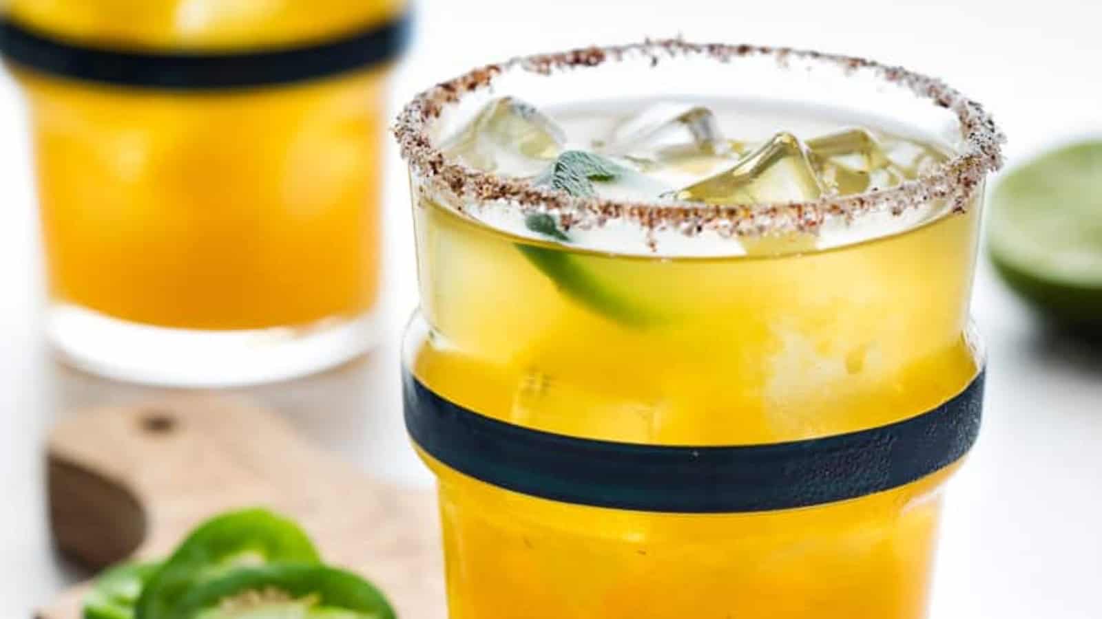 Yellow cocktail with ice, lime, and a chili-salt rim in a glass; jalapeño slices beside it.
