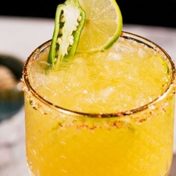 Yellow cocktail with crushed ice, chili-salt rim, lime, and jalapeño slice garnish on a marble surface.