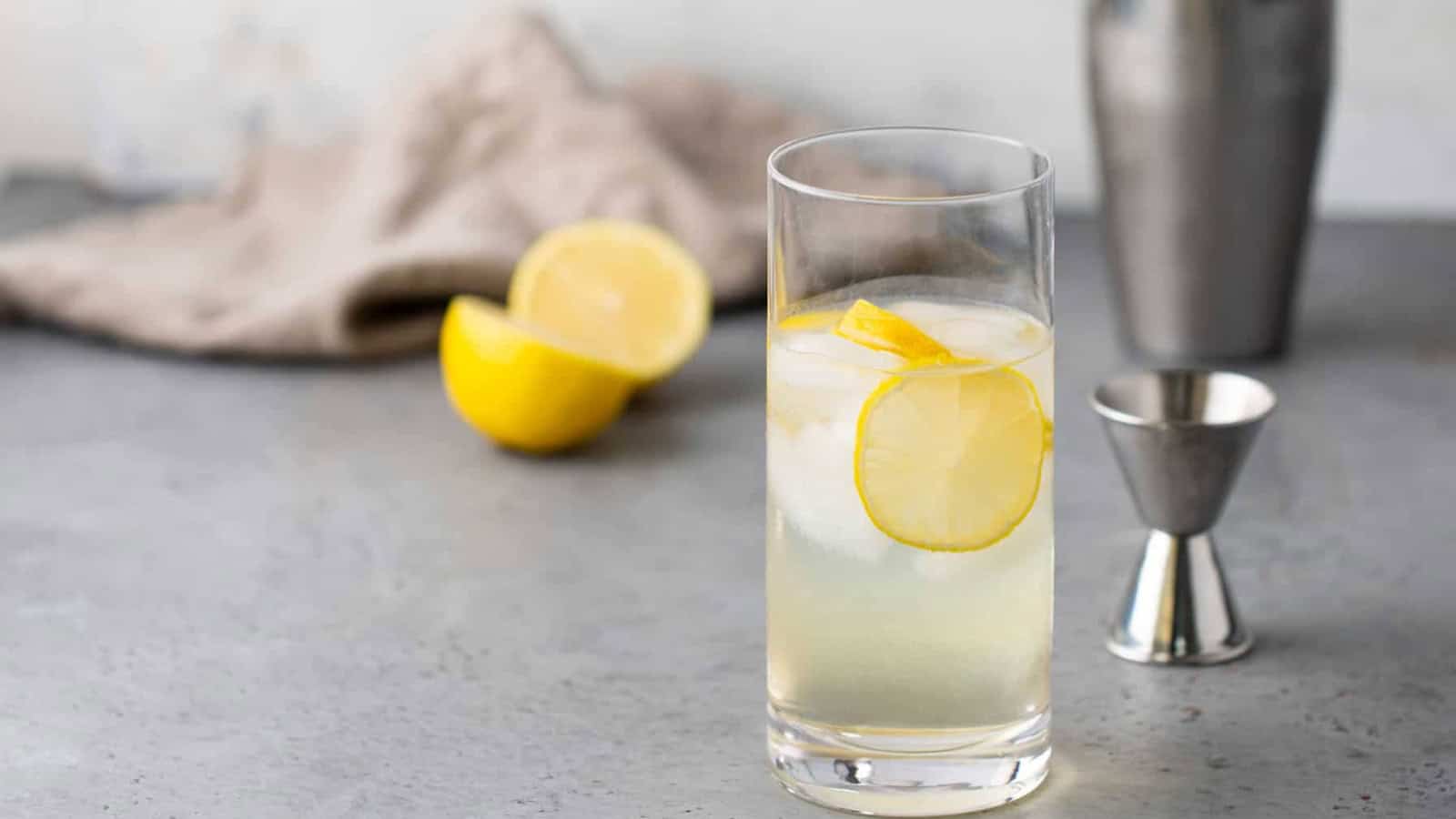 A glass of lemonade with ice and lemon slices on a gray surface, with a lemon and bar tools in the background.
