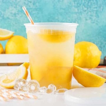 Iced lemonade in a plastic cup with a straw, surrounded by fresh lemons, ice, and striped straws.
