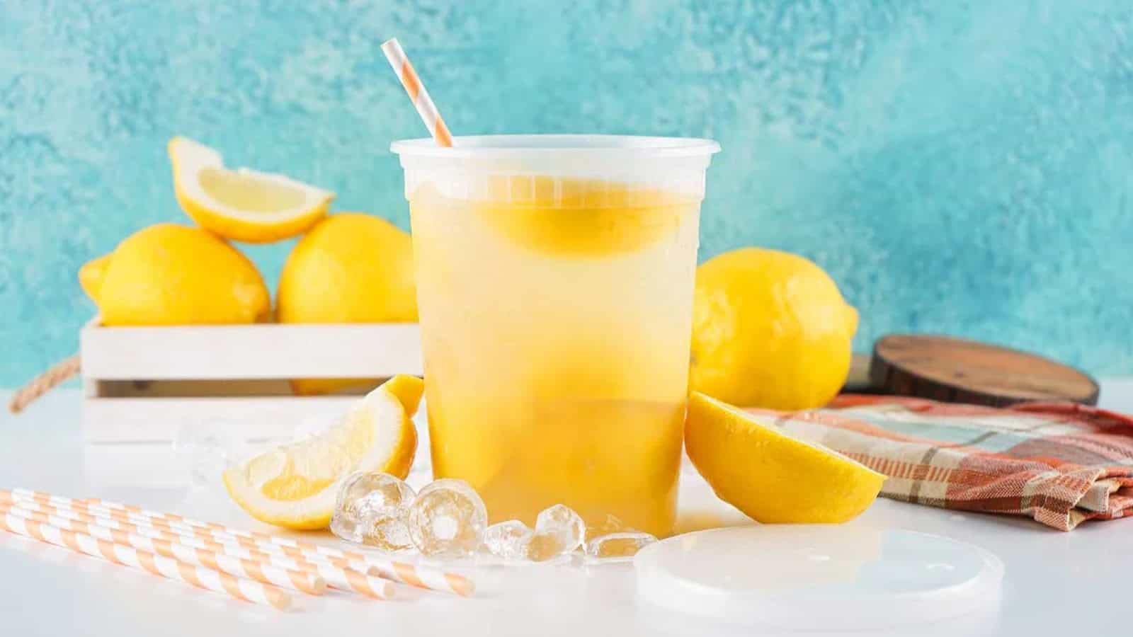 Iced lemonade in a plastic cup with a straw, surrounded by fresh lemons, ice, and striped straws.