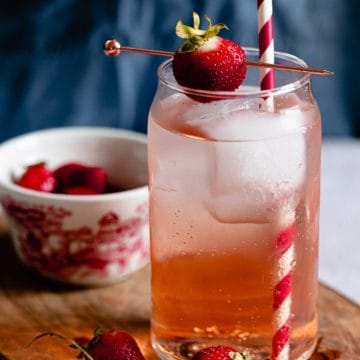 A glass of iced strawberry drink with a striped straw, garnished with a fresh strawberry. A bowl of strawberries in background.