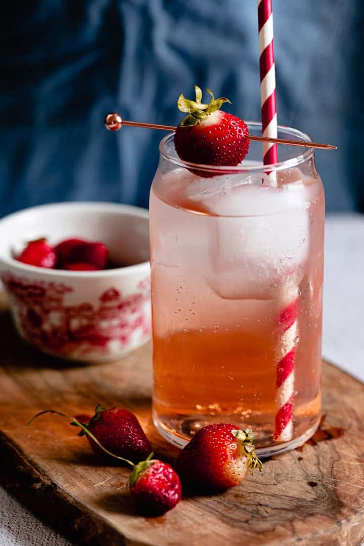 A glass of pink strawberry drink with ice, a striped straw, and a strawberry garnish on a wooden board.