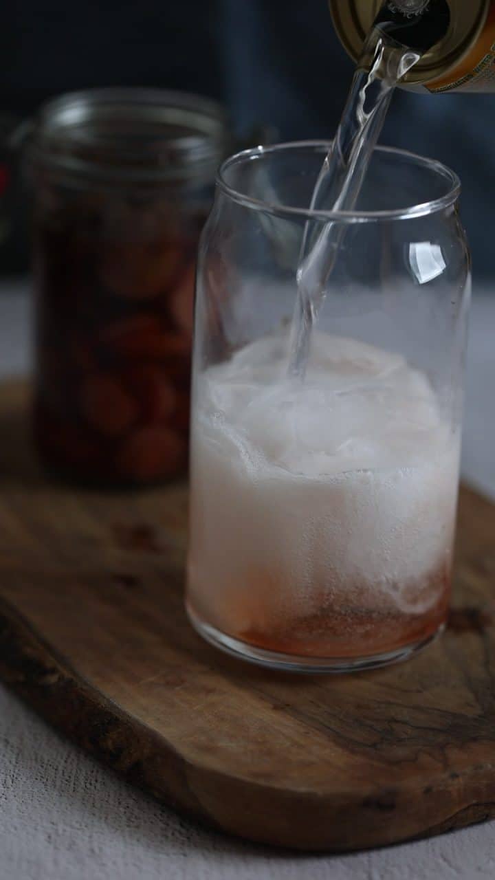 A clear drink is being poured over ice in a glass on a wooden board, with a jar of fruit in the background.