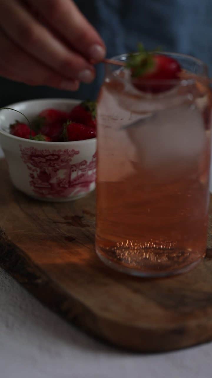 A hand places a strawberry in a glass of iced pink drink, with a bowl of strawberries beside it on a wooden board.