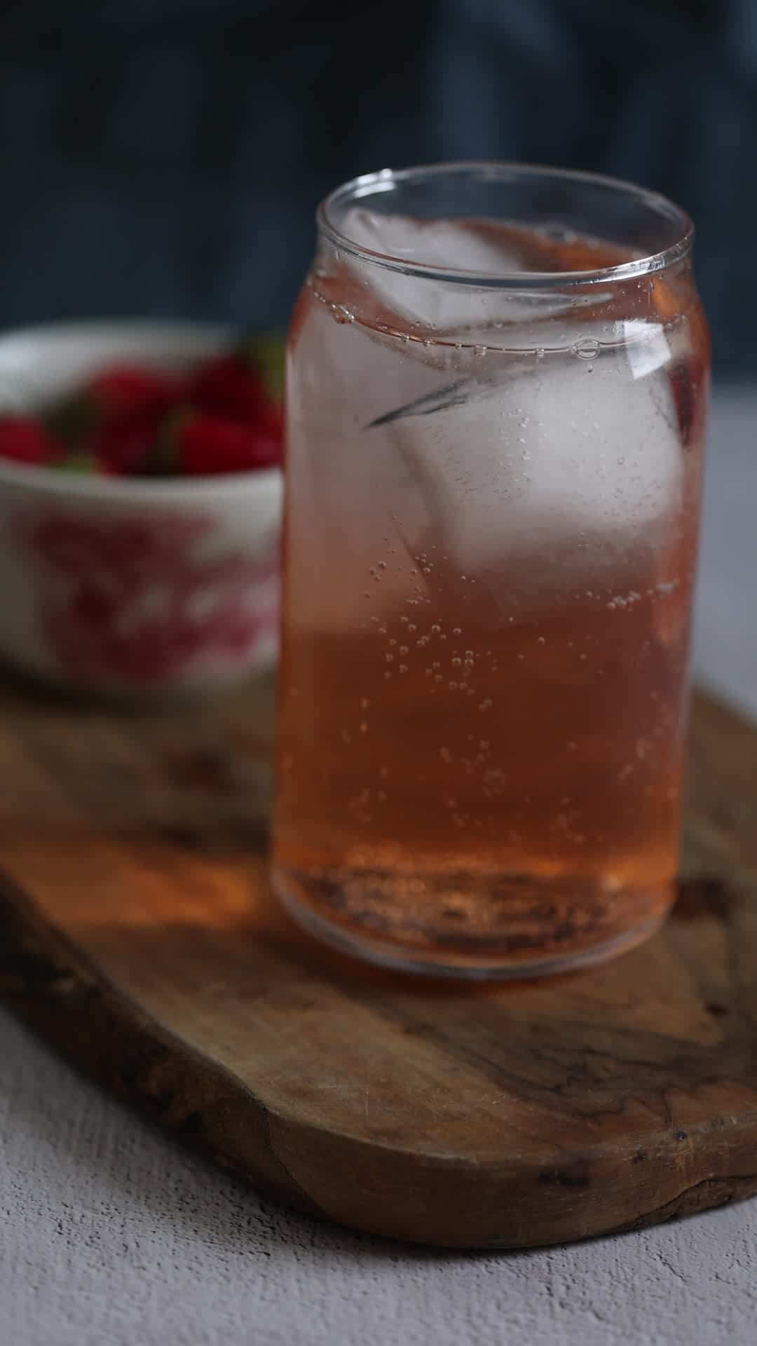 A glass of pink sparkling drink with ice sits on a wooden board, with a bowl of strawberries in the background.