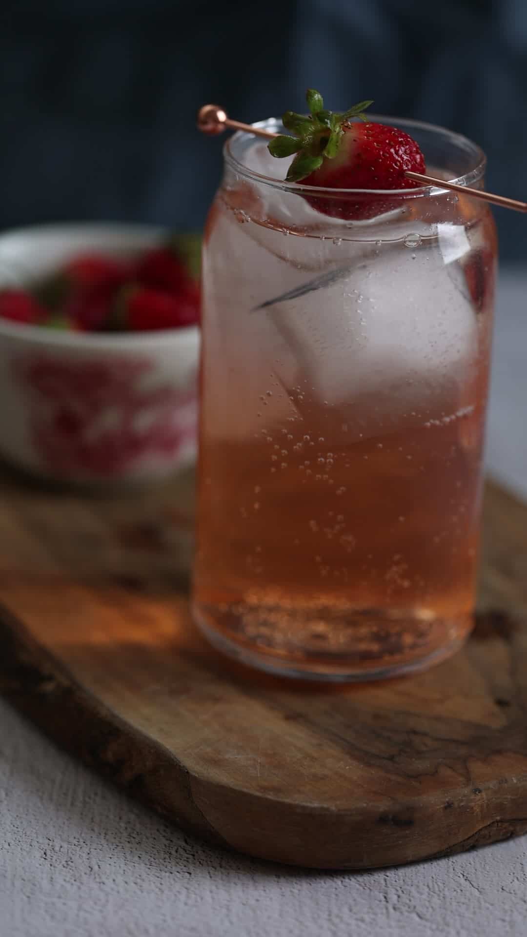 A sparkling drink with ice and a strawberry garnish, next to a bowl of strawberries on a wooden board.