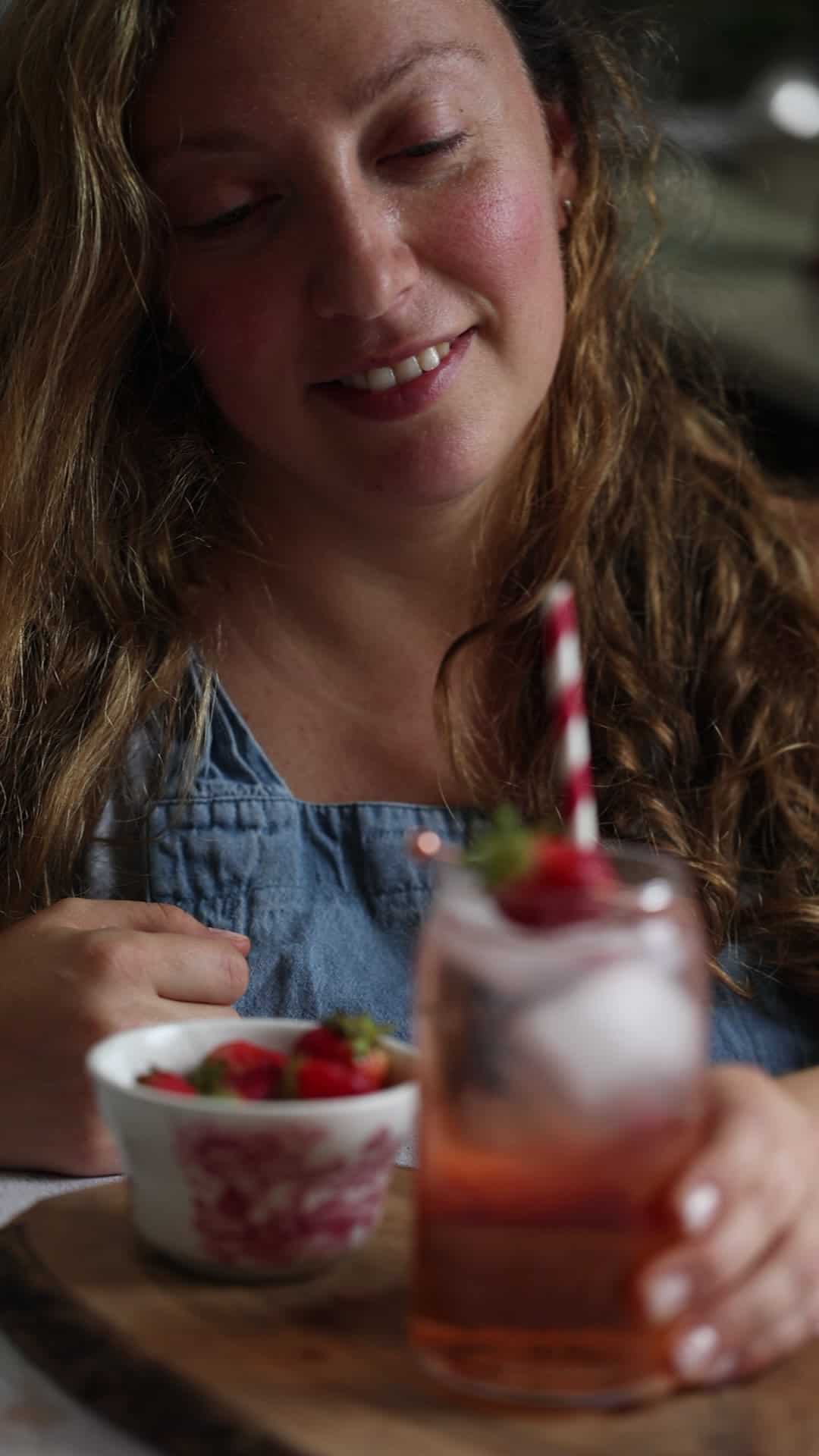 Woman smiling at a pink drink with a striped straw and a bowl of strawberries on a wooden board.