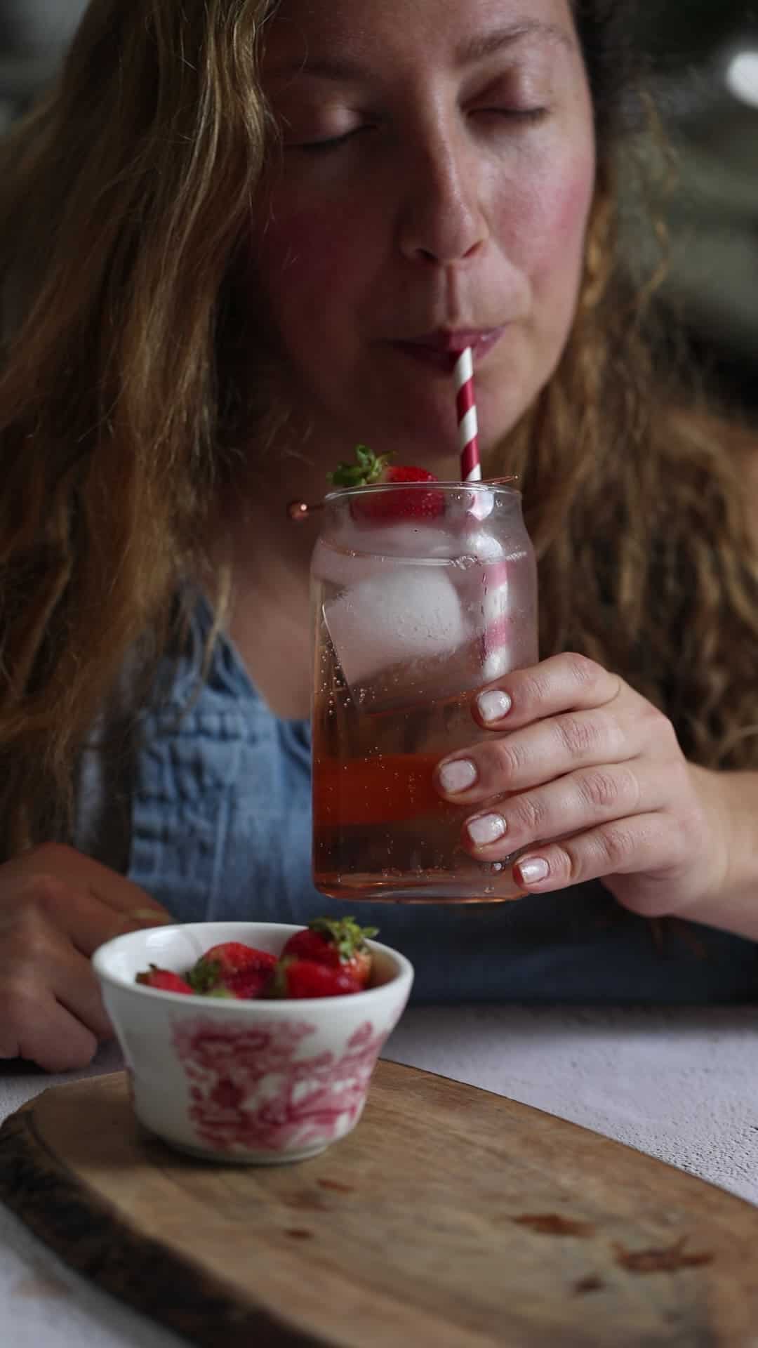 A woman drinks a strawberry beverage with ice from a jar using a striped straw.