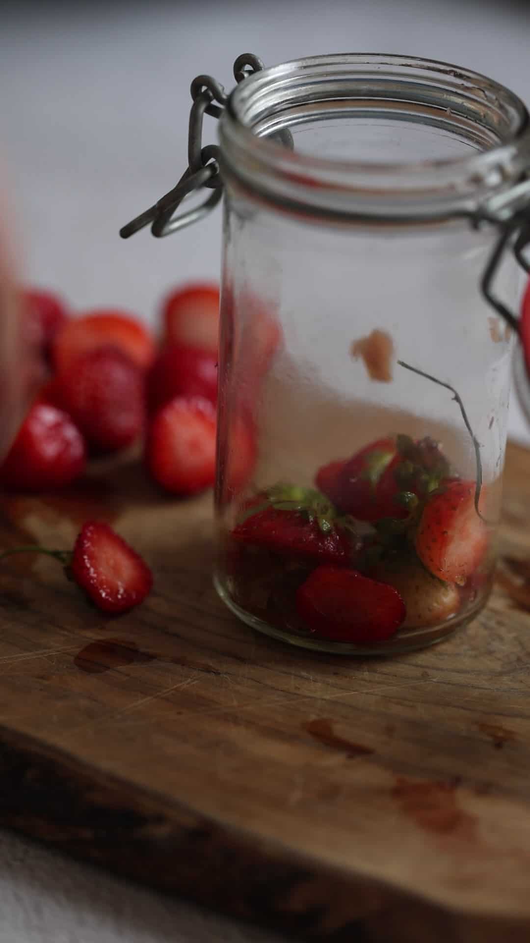 A glass jar with sliced strawberries sits on a wooden board with whole strawberries nearby.