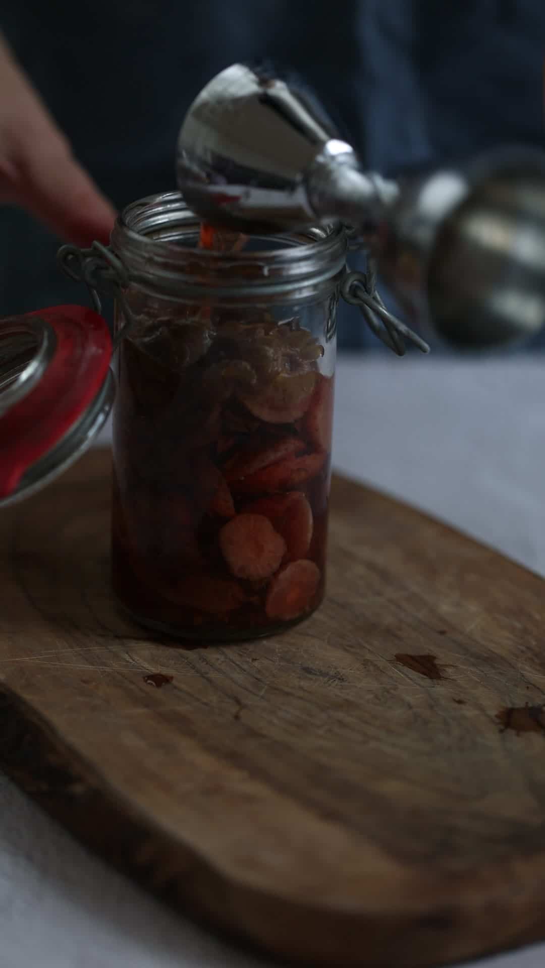 A hand pours liquid into a glass jar of sliced vegetables on a wooden board.