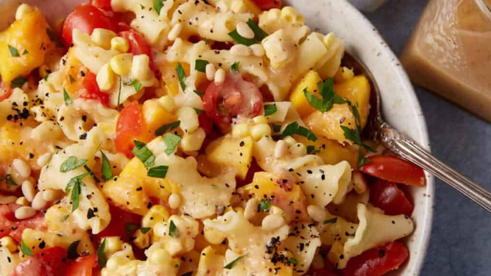 Pasta salad with tomatoes, corn, peaches, herbs, and black pepper in a white bowl with a fork.