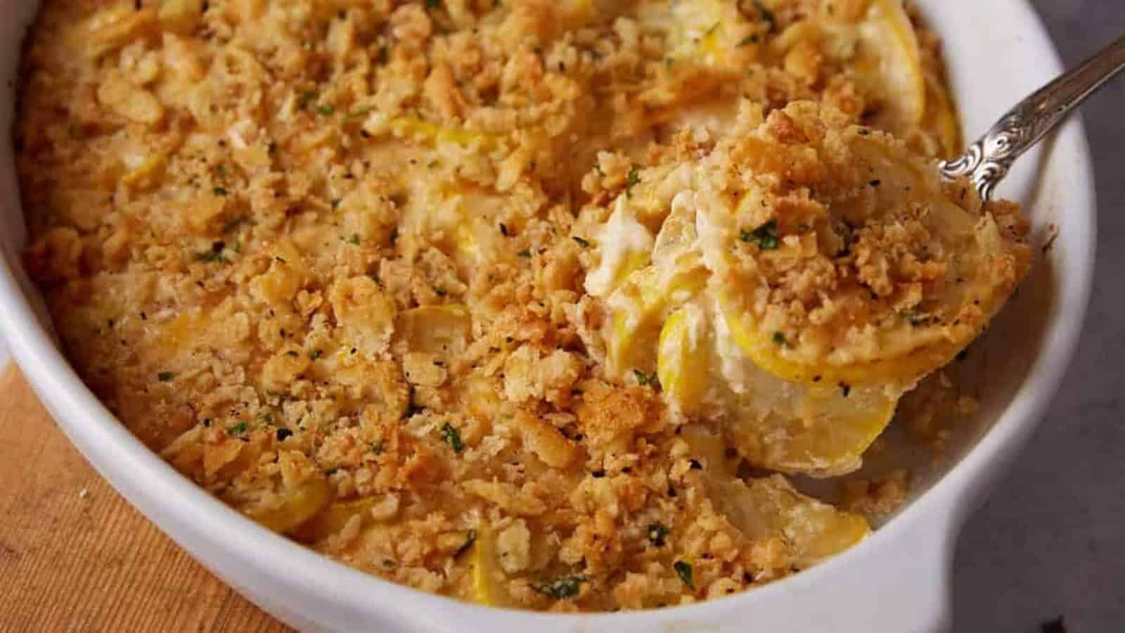 A serving spoon scoops cheesy yellow squash casserole topped with golden, crunchy crumbs from a white dish.