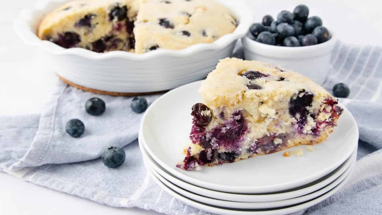 Slice of blueberry cake on stacked plates, with whole cake and bowl of blueberries in the background.
