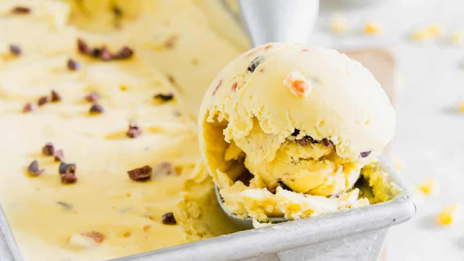 A metal scoop lifts a ball of yellow ice cream with chocolate chunks from a rectangular container.