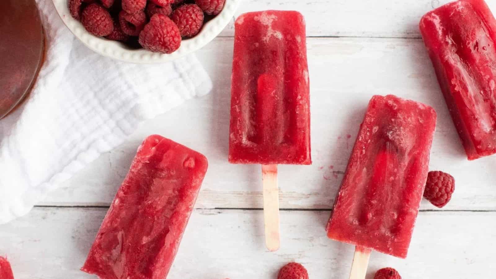 Four raspberry popsicles and fresh raspberries on a white wooden surface with a white towel.