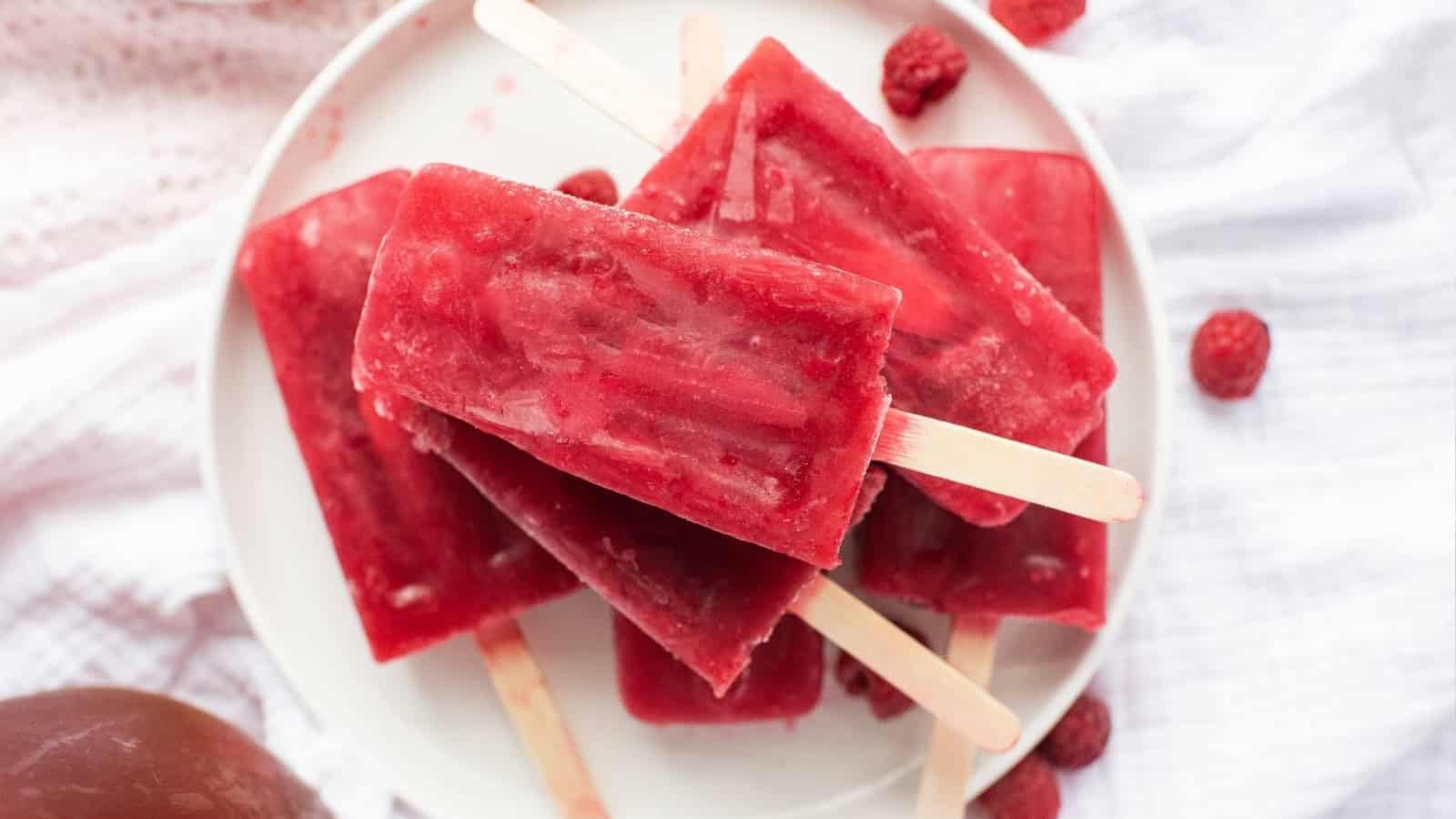 Five red berry popsicles on a white plate, surrounded by fresh raspberries on a white cloth background.