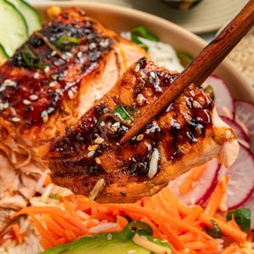 Chopsticks holding glazed grilled salmon over rice, sliced radish, cucumber, and shredded carrots.
