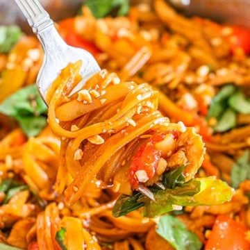 Fork holding saucy noodles with vegetables, chicken, peanuts, and basil above a pan of stir-fried noodle dish.