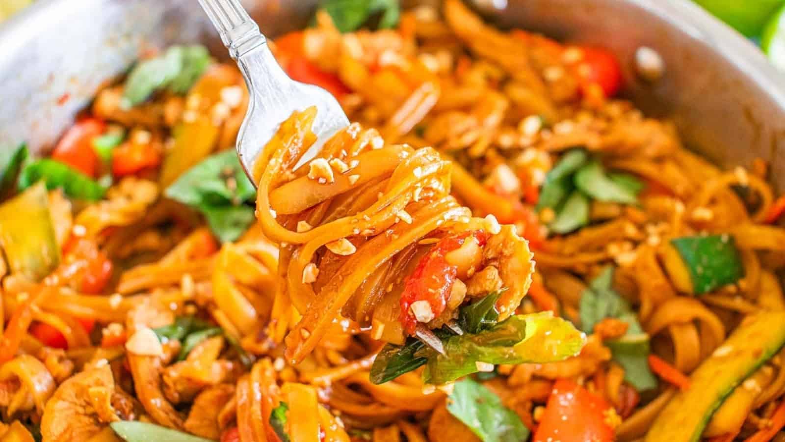 Fork holding saucy noodles with vegetables, chicken, peanuts, and basil above a pan of stir-fried noodle dish.