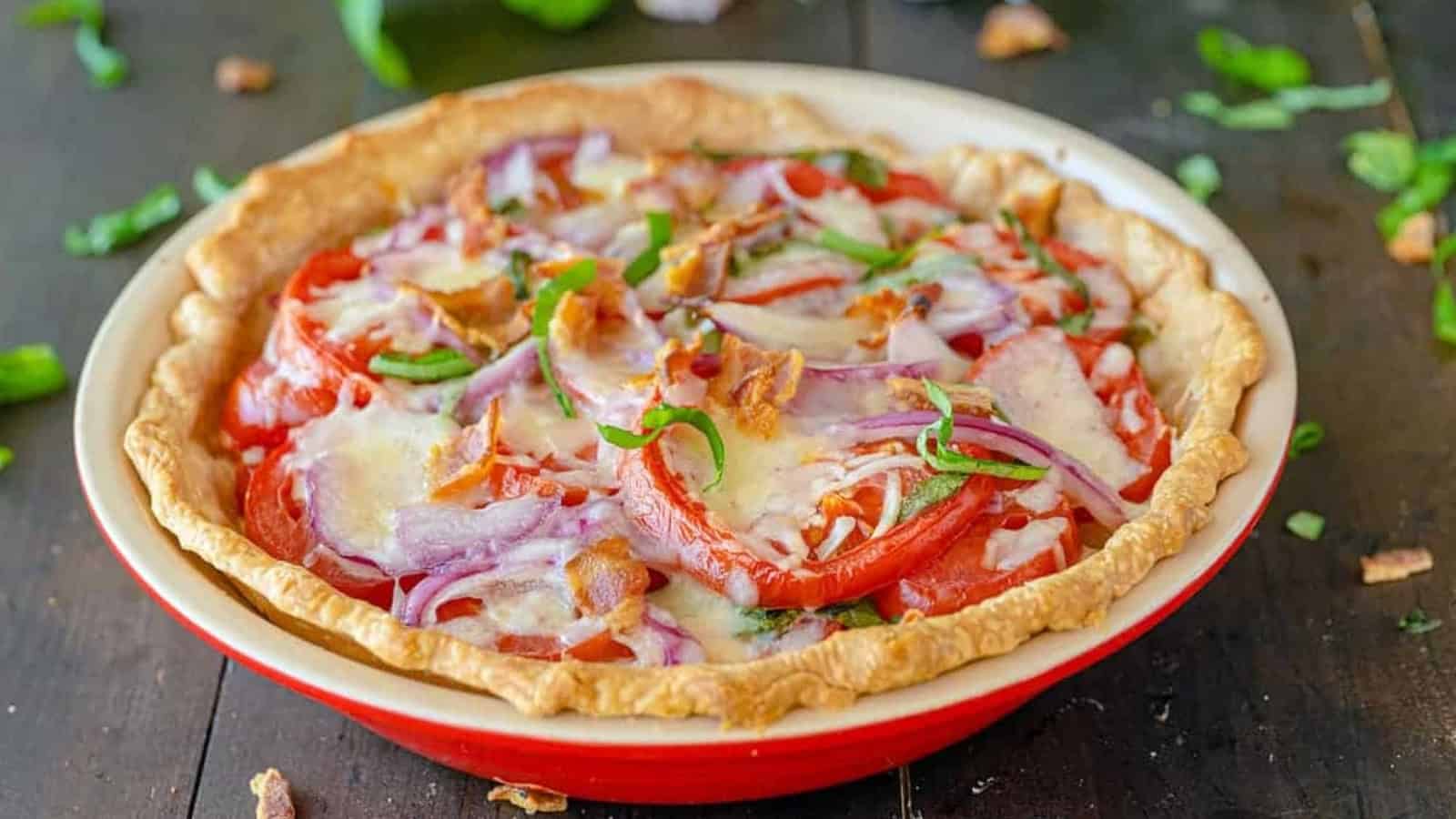 A baked pie with tomatoes, red onions, cheese, green herbs, and bacon in a red pie dish on a wooden table.